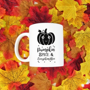 May include: White ceramic mug with a black pumpkin graphic and the text "Pumpkin Spice & Everything Nice." The mug has a white handle and is set against a backdrop of colorful autumn leaves.