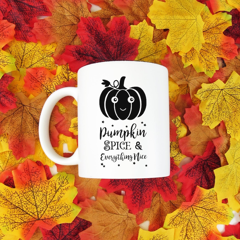 May include: White ceramic mug with a black pumpkin graphic and the text "Pumpkin Spice & Everything Nice." The mug has a white handle and is set against a backdrop of colorful autumn leaves.