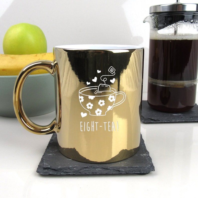 May include: Gold metallic mug with a white design of a teacup with a tea bag and the text "EIGHT-TEA!"