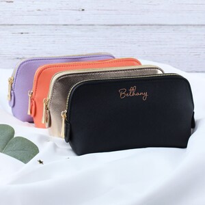 May include: Four zippered cosmetic bags in different colors: lavender, coral, gold, and black. The black bag has the name "Bethany" embroidered on it in a coral color.