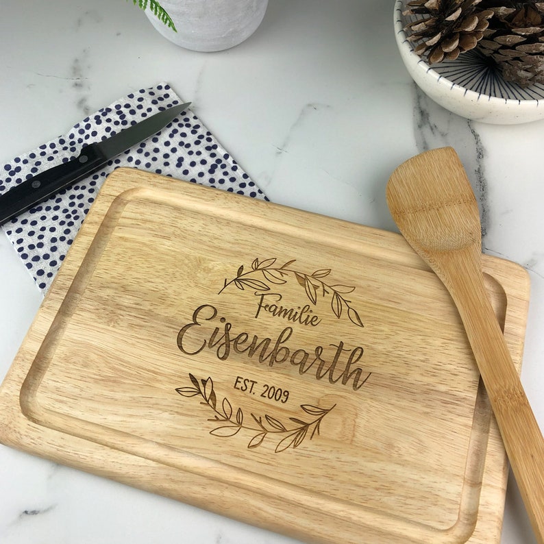 May include: A wooden cutting board with a personalized inscription that reads "Familie Eisenbarth EST. 2009" surrounded by a floral wreath design. The board is accompanied by a wooden spatula.