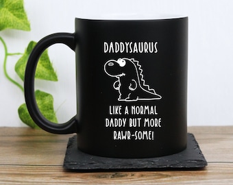 Può includere: Tazza in ceramica nera con la scritta "DADDYSAURUS" e l'illustrazione di un dinosauro. Sotto c'è la frase "LIKE A NORMAL DADDY BUT MORE RAWR-SOME!". La tazza è su un sottobicchiere quadrato nero.