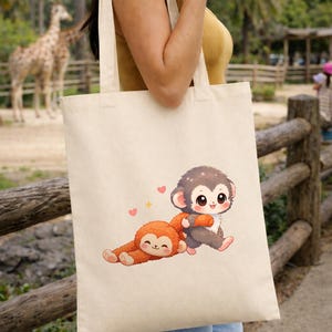 Peut inclure: Sac fourre-tout crème avec une illustration de deux singes et des coeurs. Un singe est allongé, l'autre est assis et serre un objet orange. Une girafe et une clôture en bois sont visibles en arrière-plan.