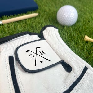 K&ouml;nnte beinhalten: Wei&szlig;er Golfhandschuh mit schwarzen Akzenten und individuellem Design. Das Design zeigt gekreuzte Golfschl&auml;ger, die Buchstaben "C" und "H" und einen Punkt. Ein wei&szlig;er Golfball und ein Holz-Tee sind ebenfalls zu sehen.