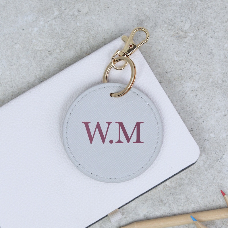 May include: A gray leather keychain with a gold metal ring and clasp. The keychain has a round, gray leather tag with the initials "W.M" in burgundy.