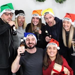 Può includere: Un gruppo di persone indossa cappelli di Babbo Natale personalizzati con nomi come Charlie, Sharon, Paige, Greg, Playdoh e Annabelle. Tengono delle bevande e sorridono per una foto.