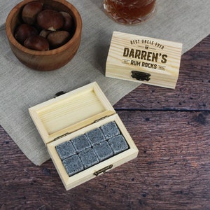May include: A wooden box with a hinged lid, containing gray stone whiskey chilling cubes. The box is engraved with the text "Best Uncle Ever Darren's Rum Rocks Est. 2004".