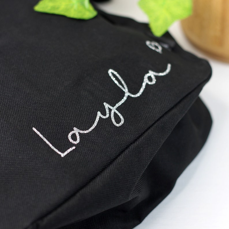 Pode incluir: Sacola de lona preta com o nome "Layla" escrito em glitter prateado.
