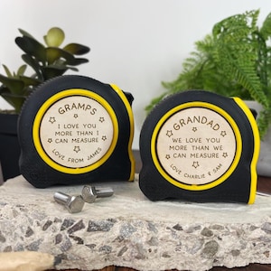 May include: Two black and yellow tape measures with personalized text. One reads "Gramps, I love you more than I can measure, love from James." The other says "Grandad, we love you more than we can measure, love Charlie & Sam."