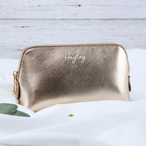 May include: A gold metallic makeup bag with a zipper closure. The bag is personalized with the name "Hayley" in white lettering.