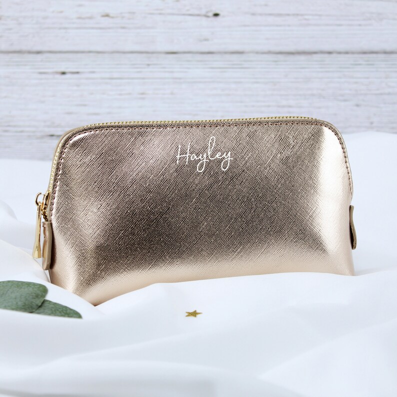 May include: A gold metallic makeup bag with a zipper closure. The bag is personalized with the name "Hayley" in white lettering.