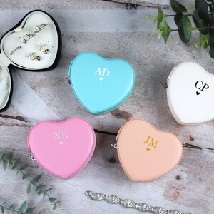 May include: A selection of heart-shaped jewellery boxes in pastel shades: light blue, pink, peach, and white. Each box is personalised with initials and a small heart. One box is open, revealing rings and a necklace.
