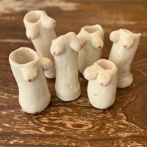 May include: A collection of five off-white ceramic vases, each sculpted in the shape of a human torso with prominent breasts featuring small, painted orange nipples. The vessels have smooth, glossy surfaces and varying heights, with open tops for holding small items.