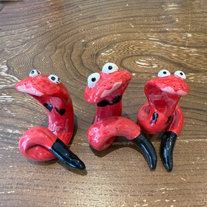 May include: Three small, glossy red ceramic snake figurines with upright heads and coiled bodies. Each snake features large white eyes with black pupils, black-tipped tails, and unique black markings on their bodies, including hearts and a bikini top.
