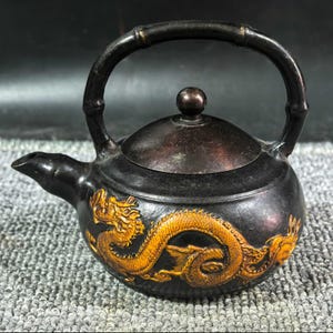 Puede incluir: Una tetera negra con un asa de estilo bambú y una tapa. La tetera presenta un diseño de dragón dorado en el lateral. El pico es pequeño y curvado. La tetera está sobre una superficie gris.