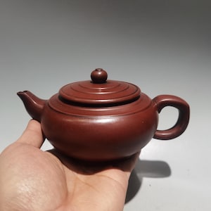 以下が含まれることがあります： 滑らかでマットな質感の暗い赤茶色の粘土製ティーポット。丸みを帯びた本体、湾曲した注ぎ口、C字型のハンドル、小さなつまみ付きの円形の蓋が特徴です。蓋には3つの同心円状のリングが装飾されています。