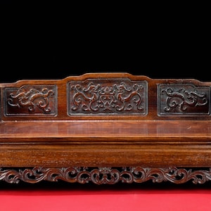 Peut inclure: Banc en bois brun foncé, finement sculpté, avec une assise rectangulaire et un dossier décoratif. Le banc présente des sculptures en relief détaillées de dragons et de motifs floraux. La base a un design en volutes. Le banc est exposé sur une surface rouge.