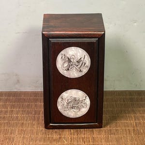May include: A dark brown wooden cabinet with two circular, decorative inlays. Each inlay features a detailed, swirling design in white and grey. The cabinet has a rectangular shape and a smooth, polished finish.