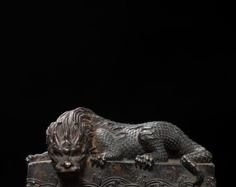 May include: A bronze-colored sculpture of a mythical creature, possibly a dragon or lion, resting on a rectangular base. The base features a wave pattern. The creature has detailed scales and a fierce expression.