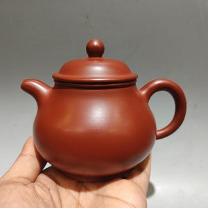 May include: A small, round, reddish-brown clay teapot with a smooth, matte finish. The teapot features a curved spout, a circular handle, and a matching lid with a small, rounded knob on top. The body is bulbous and sits low, showcasing a simple design.