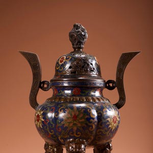 Op de afbeelding: Een sierlijke, blauw en bronzen wierookbrander met bloemen- en geometrische patronen. Het vat heeft een gewelfd deksel met een decoratieve knop en twee handvatten. De brander rust op drie sierlijke poten.