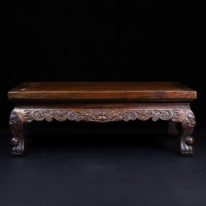 Op de afbeelding: Een rechthoekige donkerbruine houten displaystandaard met een glad, plat bovenoppervlak en een sierlijk uitgesneden rand met complexe krulpatronen. De standaard rust op vier gebogen poten die eindigen in gedetailleerde klauwvormige voeten.