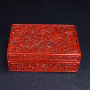 May include: A rectangular, red lacquered box with intricate floral carvings. The box features a detailed peony design on the lid and sides, showcasing a rich, textured surface. The box is a vibrant shade of red.