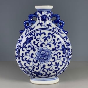May include: A blue and white ceramic flask-shaped vase. The vase has a floral design in the center and intricate patterns around the edges. It features two loop handles and a small, round opening at the top.