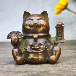 May include: A bronze Maneki Neko cat figurine, a traditional Japanese symbol of good luck. The cat is seated, holding a fan and a small sign with Japanese characters. A small pot with a frog is in front of the cat.