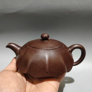 May include: A dark brown clay teapot with a rounded, petal-shaped body, a curved spout, a small circular handle, and a matching lid with a knob handle. The surface of the teapot has a matte, textured finish and a distinct scalloped design.