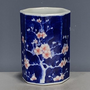 May include: A hexagonal ceramic vessel featuring a deep blue background with a white and light pink floral pattern of blossoming branches. The container has a smooth, glossy finish and a white rim, showcasing a repeating botanical design.
