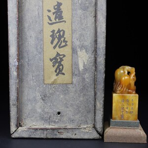 May include: An antique Chinese seal with a carved figure on top of a rectangular base. The seal is a light amber color and sits on a small, green and tan platform. A wooden box with Chinese characters is in the background.