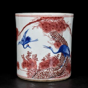 Puede incluir: Un porta pinceles cilíndrico de porcelana blanca con imágenes pintadas a mano de dos grullas azules en vuelo y posadas entre pinos rojos. El recipiente está decorado con detalles finos en esmalte rojo y azul y líneas horizontales rojas.
