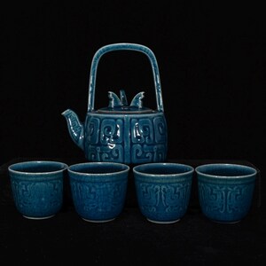 May include: A teal ceramic tea set consisting of one teapot with a high arched handle and a lid featuring small decorative protrusions, accompanied by four matching tea cups. Each piece displays a relief pattern of geometric scrollwork.