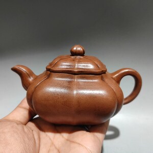 May include: A brown clay teapot with a lobed, pumpkin-like body, a matching lid with a small knob handle, a curved spout, and a rounded handle. The surface has a matte, earthy texture with visible vertical ridges defining the segments.
