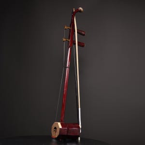 Puede incluir: Un erhu chino tradicional, un instrumento musical de arco de dos cuerdas. El erhu presenta un cuerpo y mástil de madera marrón rojizo oscuro, con un arco apoyado contra las cuerdas. El instrumento se encuentra sobre un fondo oscuro.
