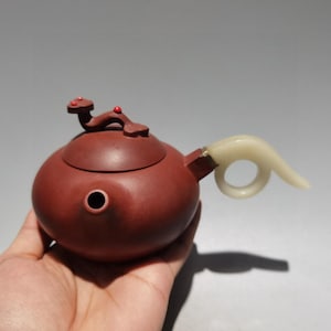 Può includere: Una teiera in argilla marrone rossastro scuro con un corpo liscio e arrotondato e un beccuccio corto. Il coperchio è sormontato da una scultura di una creatura decorata con due piccole perline rosse. Un manico curvo color bianco sporco, simile alla giada, è fissato lateralmente.
