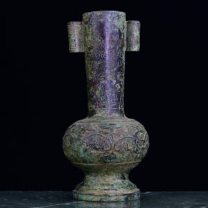Puede incluir: Un jarrón cilíndrico alto de tono bronce con una pátina desgastada y moteada en verde y marrón oscuro. El cuello presenta columnas verticales de caracteres grabados, mientras que el cuerpo bulboso muestra relieves simétricos.