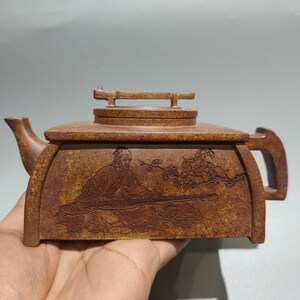May include: Brown rectangular teapot with a lid and handle. The teapot features an engraved image of a seated figure and floral elements. The spout and handle are curved. The teapot is held in a hand.