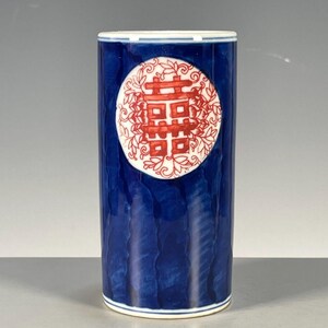 May include: A cylindrical ceramic vase featuring a deep blue glazed body with a textured, wavy surface. A prominent white circular medallion on the front displays a red double happiness symbol surrounded by intricate red floral line art patterns.