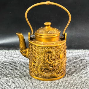 Könnte beinhalten: Eine goldfarbene Teekanne mit Henkel und Deckel. Der Körper der Teekanne ist aufwendig mit einem Drachendesign verziert. Der Ausguss ist gebogen und der Griff wölbt sich über die Oberseite. Die Teekanne ist wahrscheinlich aus Metall und hat ein dekoratives Aussehen.
