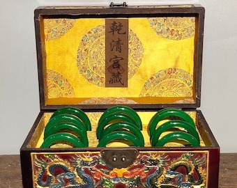 Puede incluir: Una caja de madera con un interior dorado y rojo y un diseño de dragón en la parte delantera. La caja está abierta y contiene varios brazaletes de jade verde. La tapa de la caja tiene una etiqueta con caracteres chinos que dicen "乾隆年制".