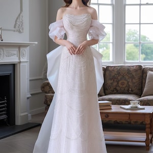 Op de afbeelding: Een witte trouwjurk met een off-the-shoulder ontwerp, versierd met kralen op het lijfje en een zwierige rok. De jurk heeft doorschijnende, gerimpelde mouwen en een grote strik op de rug. De bruid staat in een lichte kamer.