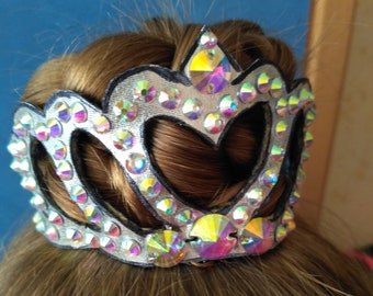 May include: A silver tiara with iridescent rhinestones, set against brown hair. The crown features a heart-shaped cutout design and is adorned with multiple colorful gems. The tiara is likely for a special occasion or performance.