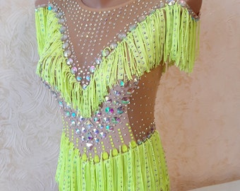 May include: A neon yellow leotard with fringe detailing and sparkling rhinestones. The garment features a nude mesh overlay on the shoulders and torso, and is designed for dance or gymnastics performances.