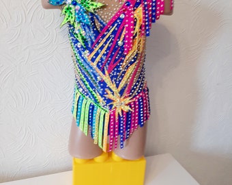 May include: A colourful gymnastics leotard with intricate detailing. The leotard is predominantly blue, pink, and yellow, with rhinestone and ribbon embellishments. A large floral applique is on the shoulder, adding a decorative touch to the athletic garment.