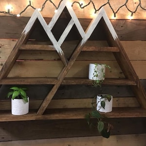 Puede incluir: Una estantería de madera con forma de montaña con tres estantes. La estantería está pintada de blanco en la parte superior y tiene un acabado marrón rústico. Dos pequeñas plantas en maceta están en los estantes.