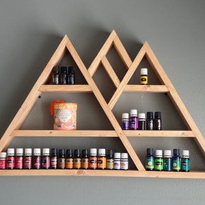 Puede incluir: Una estantería triangular de madera con múltiples estantes, que muestra una variedad de botellas de aceites esenciales en varios colores, incluyendo morado, verde, naranja y marrón. La estantería está diseñada para parecer una cordillera.