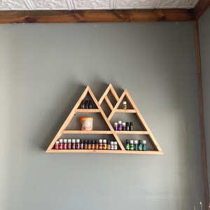 May include: A wooden triangular shelf unit with multiple shelves, displaying a variety of essential oil bottles and a candle. The shelf is mounted on a gray wall.
