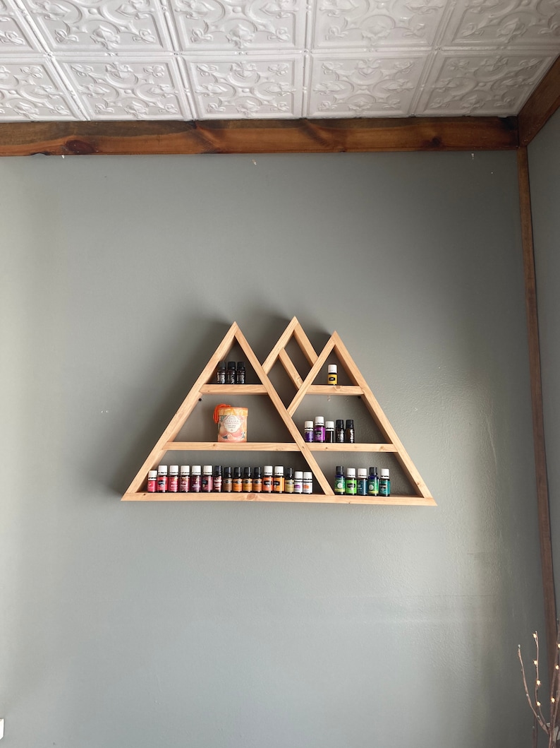 May include: A wooden triangular shelf unit with multiple shelves, displaying a variety of essential oil bottles and a candle. The shelf is mounted on a gray wall.